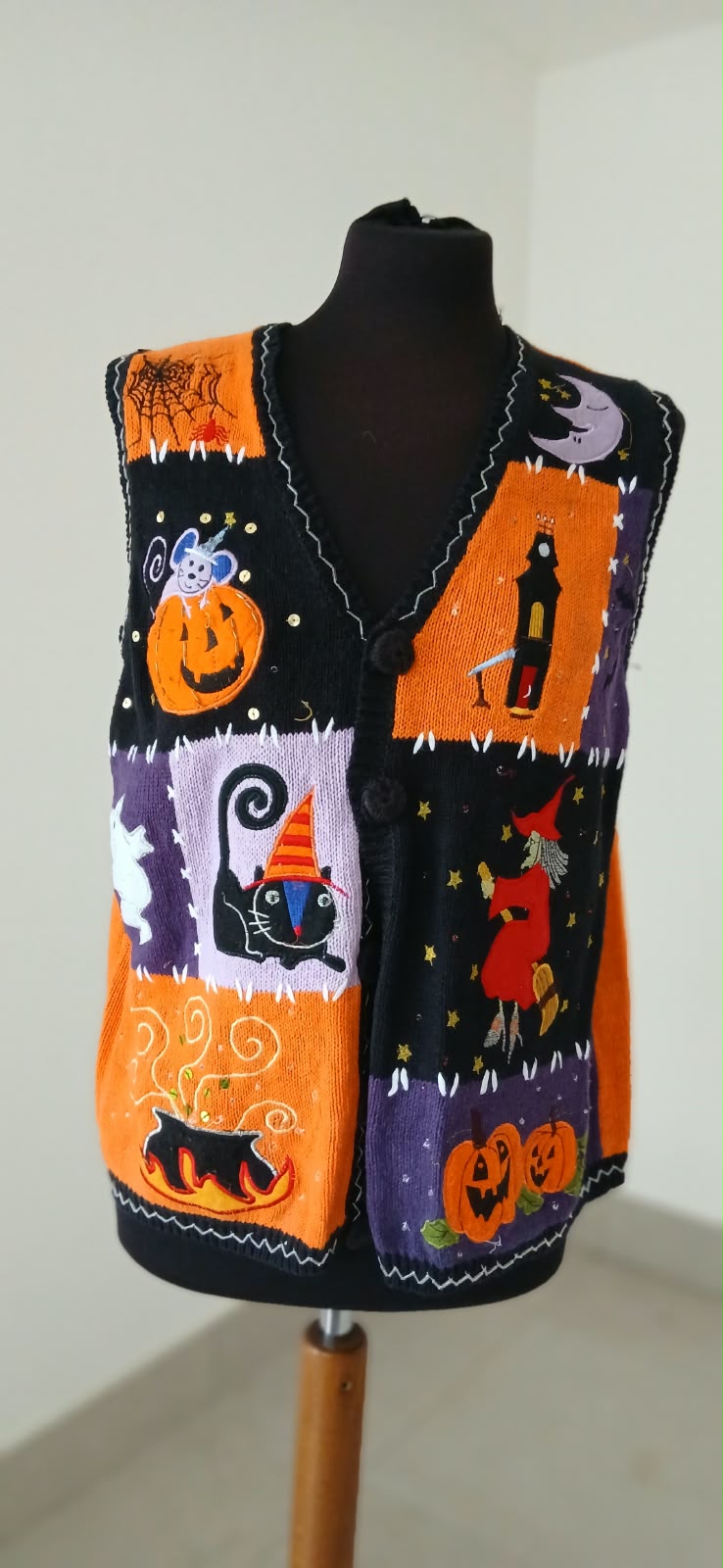 A sleeveless black knit vest with orange trim and colorful Halloween-themed patchwork panels, displayed on a black mannequin torso against a plain indoor wall.