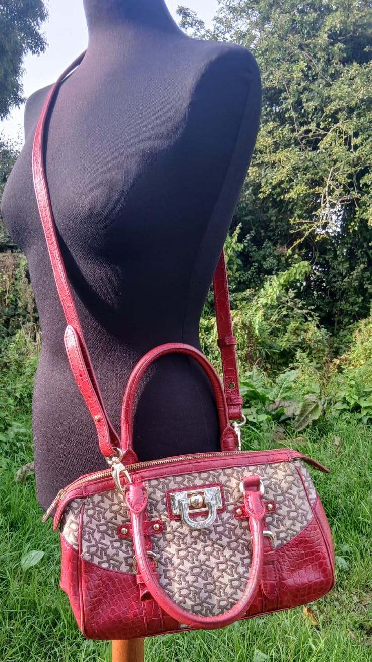 A red and beige monogram handbag displayed on a black mannequin torso outdoors, with green grass and trees in the background.