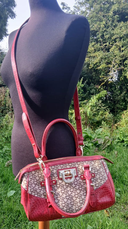 A red and beige monogram handbag displayed on a black mannequin torso outdoors, with green grass and trees in the background.
