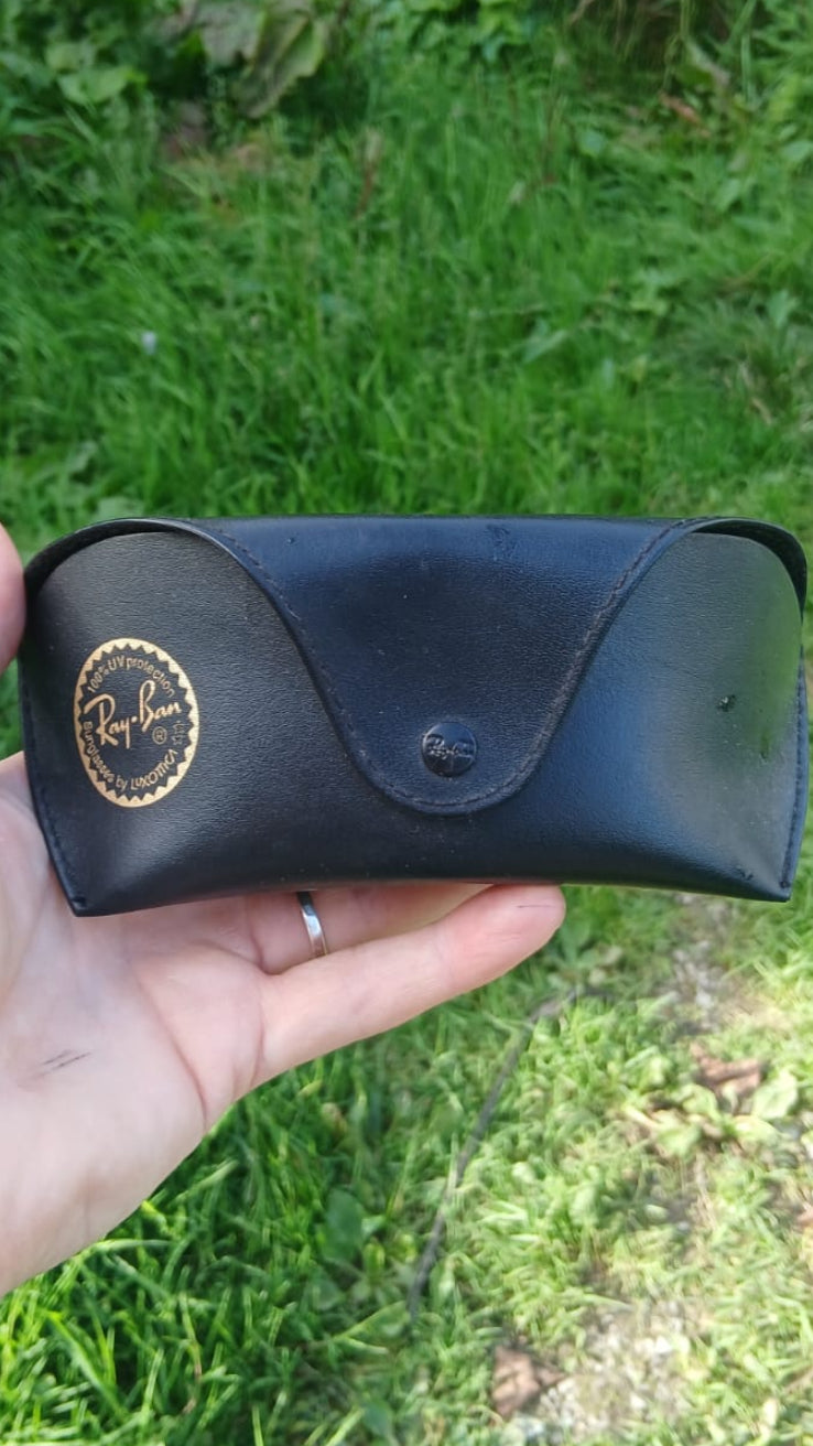 A black leather Ray-Ban glasses case featuring a gold circular Ray-Ban logo on the left and a snap button closure.