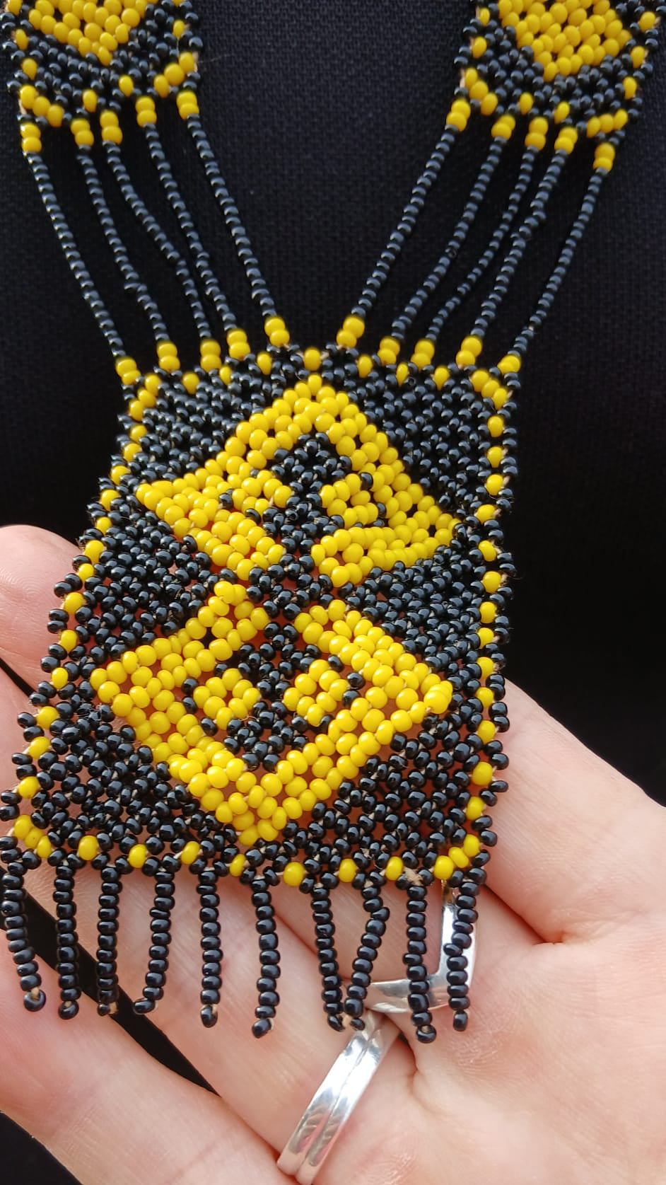 A close-up of a beaded necklace with a large geometric pendant. The necklace features black and yellow seed beads, with a central diamond pattern in yellow and black on a black background.