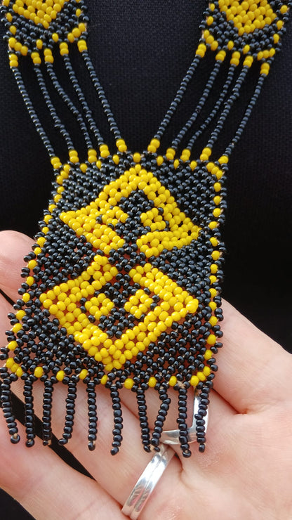 A close-up of a beaded necklace with a large geometric pendant. The necklace features black and yellow seed beads, with a central diamond pattern in yellow and black on a black background.