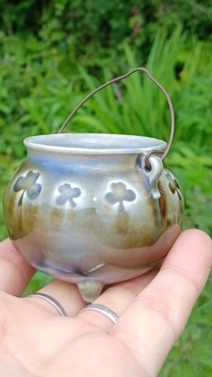 A close up of a handmade ceramic cauldron with a handle from Wade Ceramics, featuring a green and blue glaze and raised shamrock relief details.