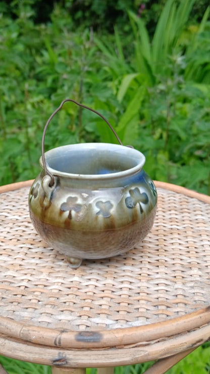 A small handmade ceramic cauldron with a handle from Wade Ceramics, featuring a green and blue glaze and raised shamrock relief details.