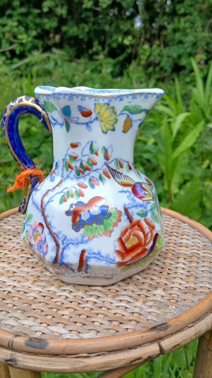A hand-painted ceramic jug with a multicolor floral and bird design and a blue handle.