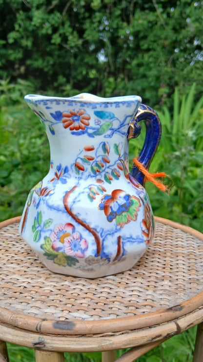 A white english fine earthware pitcher decorated with colorful floral motifs, featuring a dark blue handle.