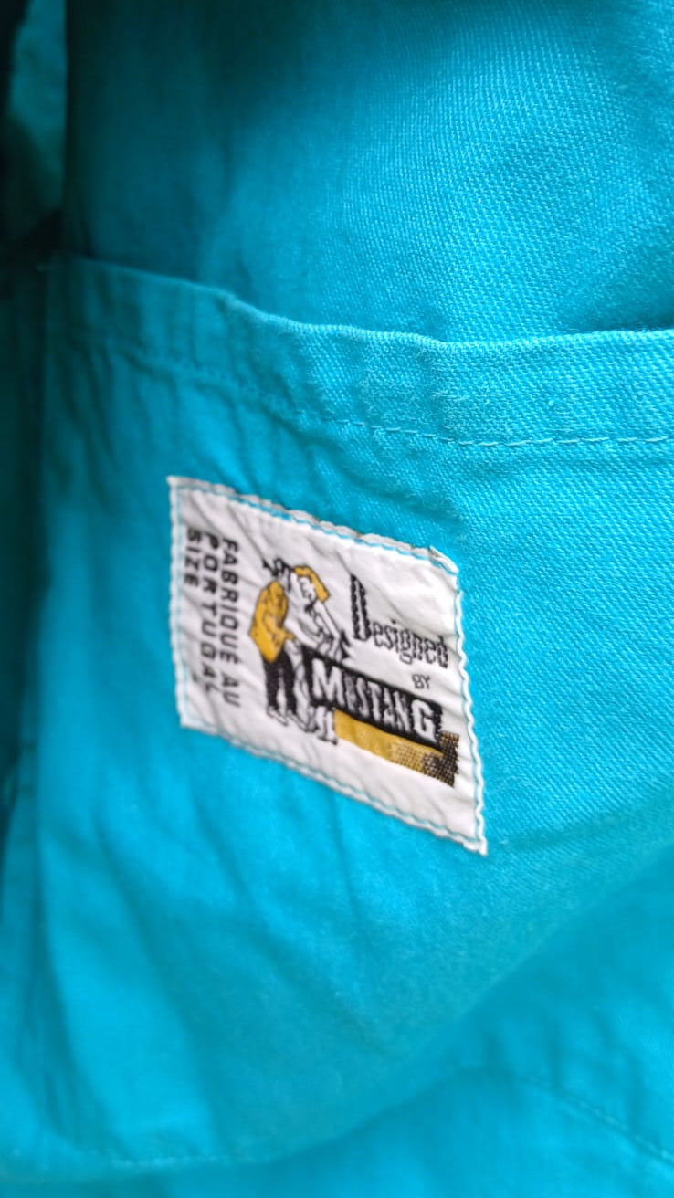 Close-up of a turquoise blue fabric garment with a white sewn-in brand patch reading Designed by Mustang.