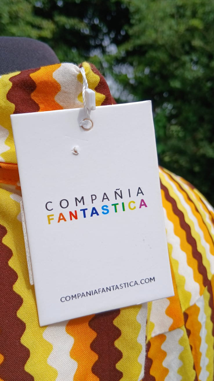 A white paper hangtag with the text 'COMPAÑIA FANTASTICA' in black and rainbow-colored letters, attached to a yellow, orange, brown, and white zigzag-patterned midi skirt.