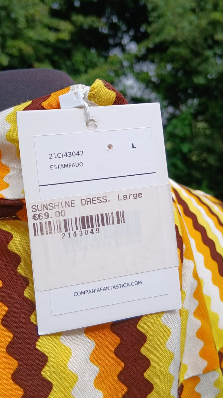 A white garment tag for a 'SUNSHINE DRESS. Large' size L, held up against a colorful yellow, brown, and white wavy patterned midi skirt collar.
