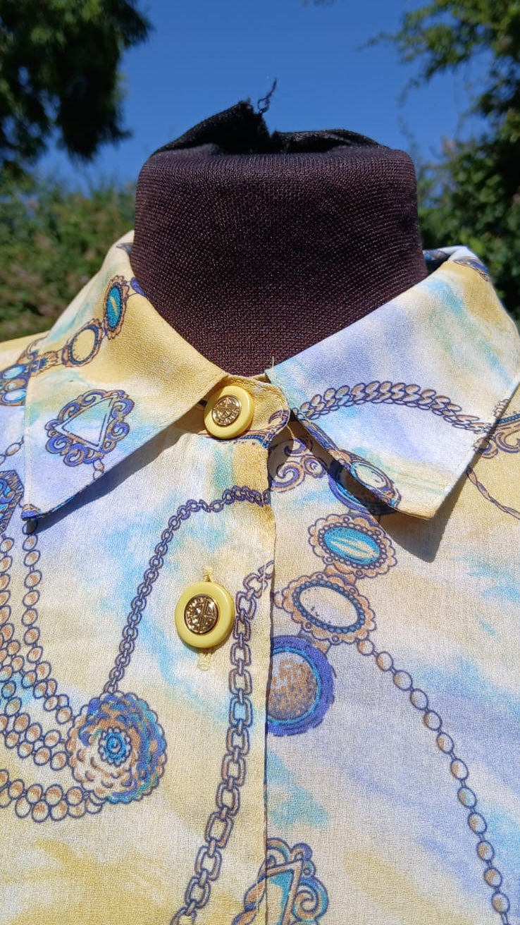 A close-up of a collared button-front shirt with a pale yellow base and a blue jewelry chain-link pattern and yellow buttons with gold accent.