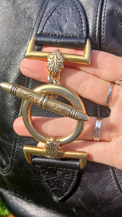 A close-up of a black leather bag strap with a large ornate toggle clasp. The clasp is made of gold-toned metal with engraved details and a circular ring element.